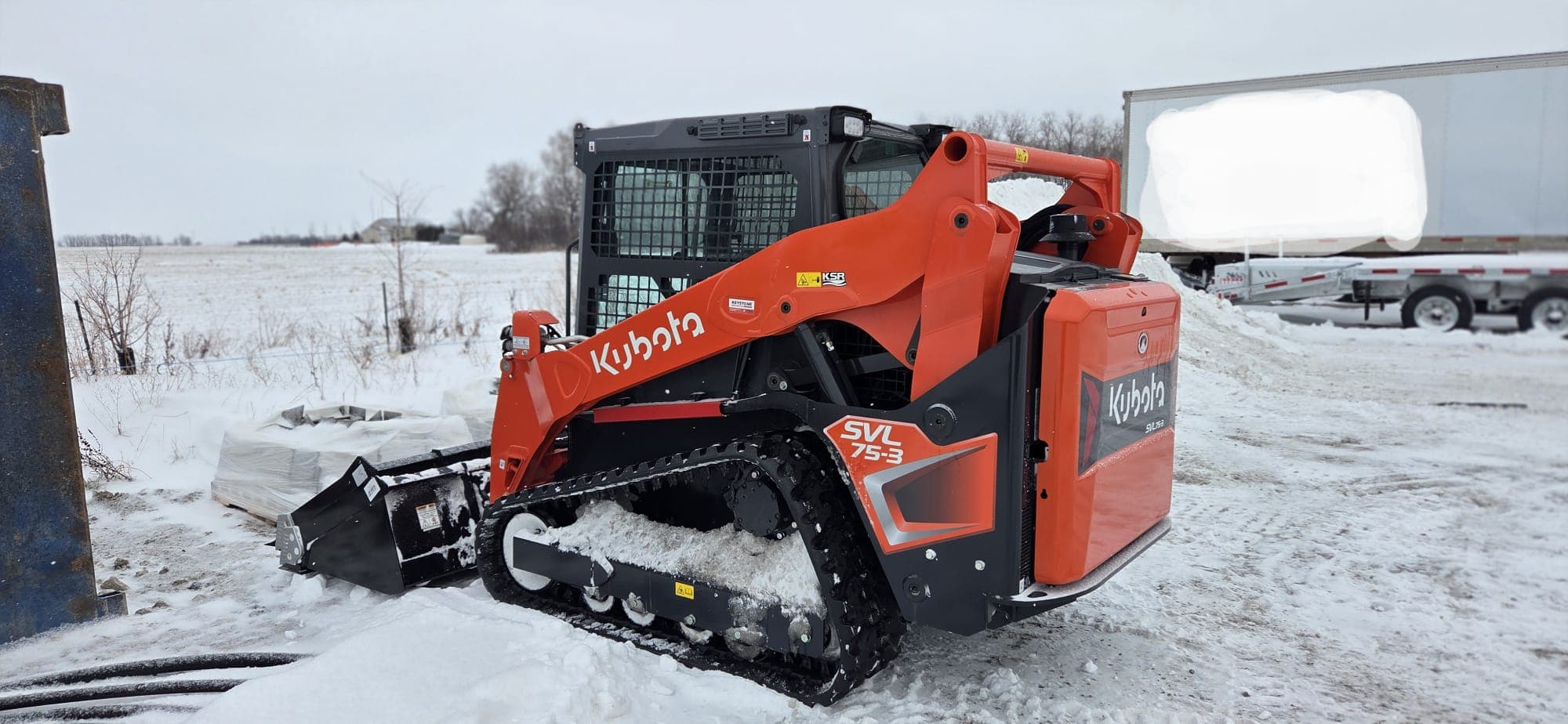 Orange Kubota SVL 75-3 track loader with a bucket attachment parked in a snowy field.