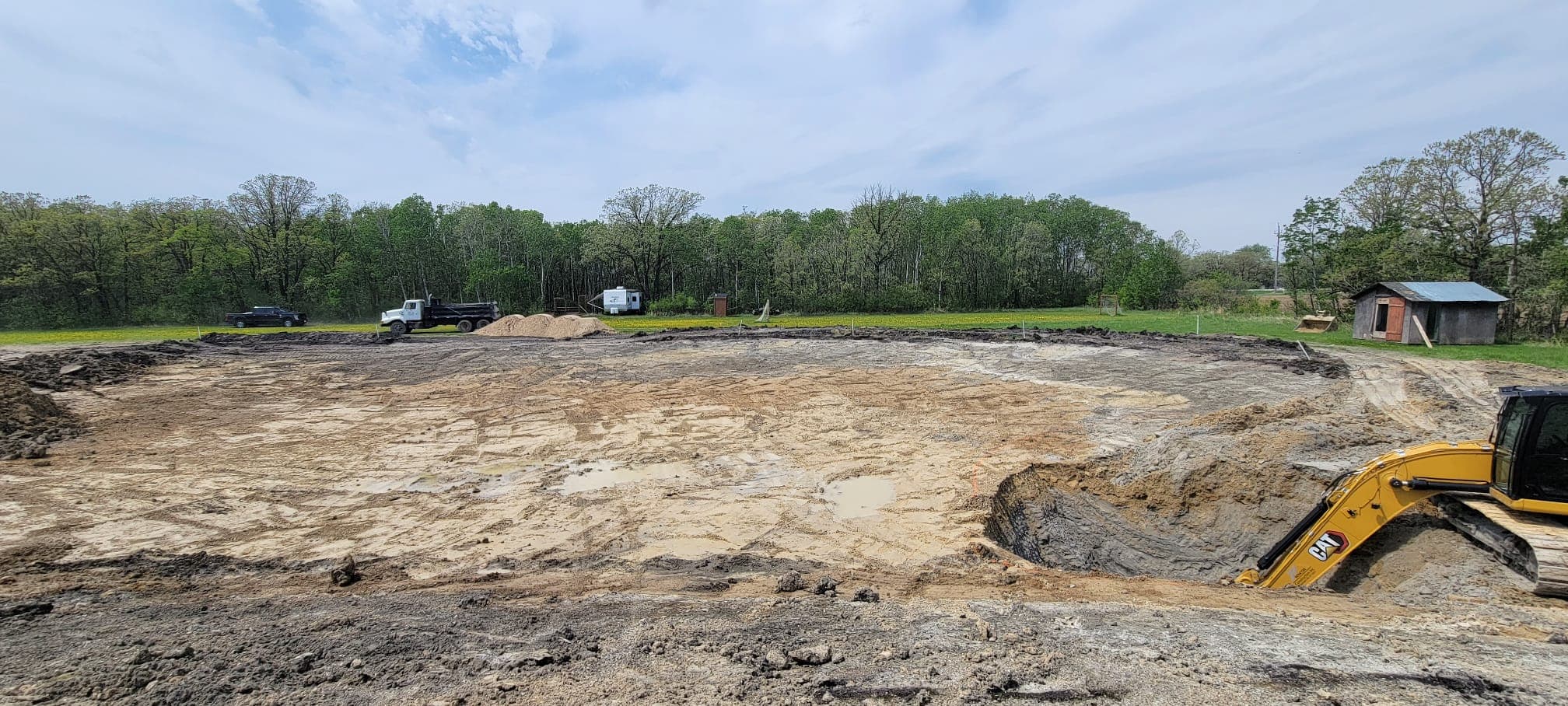 Yellow excavator digging a large pit in a muddy construction site surrounded by green trees.