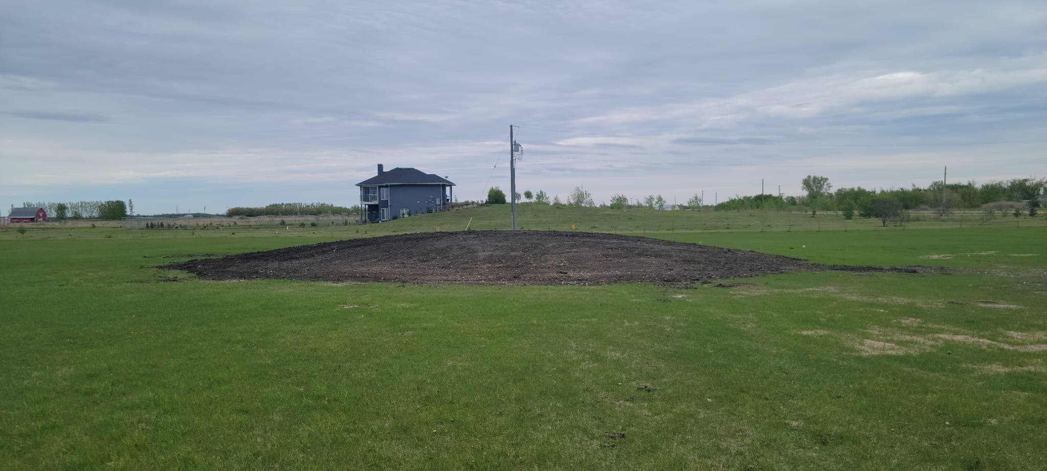 Large mound of dark soil in a green field with a blue house and clouds.